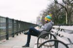 Lifestyle image of a man sitting on a bench. The colours are monotone and the vibrant mustard hat pops in the picture.