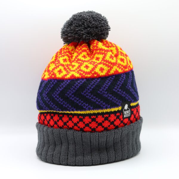 The Ultra Wonky hat is knitted with a grey pompom and turn up. Sandwiched between is the triple pattern area knitted in vibrant red and yellow with navy and purple contrasting features.