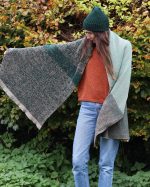 Woman modelling a green and fawn knitted blanket scarf, fanned out her right arm to show the product.