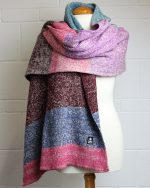 Image of a model wearing a blanket scarf, knitted in wool using cream, pink, burgundy and blue hues.