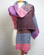 Image of a model wearing a knitted wool blanket scarf. The colours are burgundy, pink and blue hues on a cream base.
