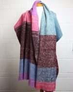 Image of a mannequin wearing a blanket scarf draped over the shoulders. The scarf design has blocks of colours including blue, pink and burgundy set onto a cream base.