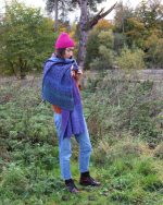 Image of a model outdoors wearing a pink beanie and a purple knitted blanket scarf.