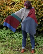 Image of a model wearing a blanket scarf outdoors. The colours in the scarf are blue, red, grey, green knit in block colours.